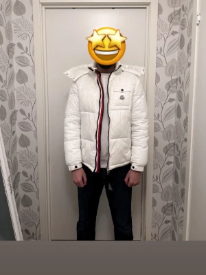 Vit pufferjacka från Moncler - Säljer en vit pufferjacka från Moncler med avtagbar huva och coola röd-blå detaljer längst dragkedjan. Jackan har en bröstficka med knapp och Moncler-logga, samt två sidofickor. Perfekt för kalla dagar och riktigt snygg stil.