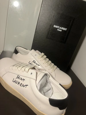 Saint Laurent skor - Säljer ett par snygga vita sneakers från Saint Laurent med handskriven logga på sidan. Skorna har svart detalj vid hälen, beige platt sula och klassisk snörning. Tillverkade i skinn för en lyxig känsla och kommer med originalförpackning.