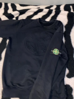 Svart sweatshirt från Stone Island - Svart sweatshirt från Stone Island med stort broderat logomärke på bröstet och klassisk patch på ena ärmen. Tröjan har rund halsringning och långa ärmar. Perfekt för dig som gillar streetwear och vill ha en stilren look. Den skulle passa till alla ”XS” men funkar om du är 170 till 175 cm lång 