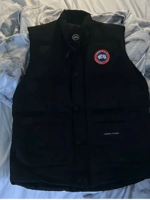 Svart dunväst från Canada Goose - Svart dunväst från Canada Goose med klassisk logotyp på bröstet och hög krage. Västen har praktiska fickor framtill och diskret knappstängning. Materialet är slitstark polyester och dunfyllning som håller värmen. Perfekt för kyliga dagar och har en stilren look.