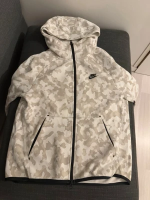 Nike camo mjukisbyxor med dragkedja - Säljer ett par mjukisbyxor från Nike i beige och vit camouflage-mönster. Byxorna har svarta snören i midjan, svarta dragkedjor på fickorna och Nike-logga på benet. Materialet är mjukt och skönt, perfekt för chill eller träning. Byxorna är storlek S! men passar både M.