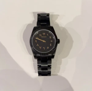 Svart seiko mod, automat, arabisk siffror - Bilderna visar en svart Seiko-klocka med arabiska siffror på urtavlan och metallarmband, ingen väst syns.