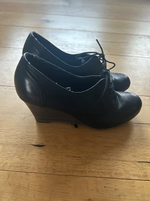 Svarta kilklackskor från Tamaris - Snygga svarta skor från Tamaris med kilklack och snörning framtill. Skorna har rund tå och är gjorda i skinn med en stabil plattformsula. Perfekta för dig som vill ha både stil och komfort i vardagen.