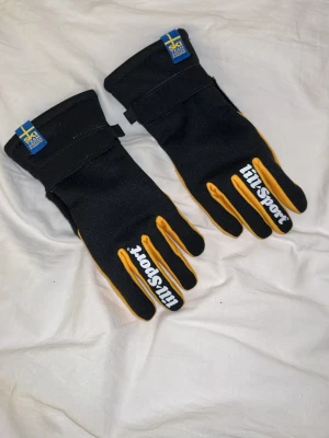 Svarta och gula Tillsport handskar - Svarta handskar från Tillsport med gula detaljer längs fingrarna och vit logga på ovansidan. Handskarna har svensk flagga och SAI Ski Team Sweden-märke vid handleden. Perfekta för kalla dagar och utomhusaktiviteter.