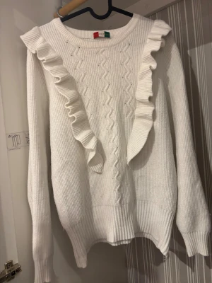 Vit stickad tröja med volang från Donna Di Classe - Säljer en vit stickad tröja från Donna Di Classe med snygga volangdetaljer längs axlarna och fram. Tröjan har ett klassiskt kabelstickat mönster på framsidan, rund hals och långa ärmar. Perfekt för dig som gillar en feminin och trendig look.