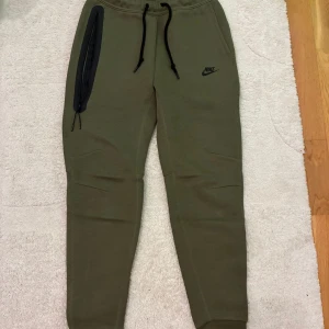 Nike Tech mjukisbyxor -  olivgröna mjukisbyxor från Nike Tech med svarta snören i midjan och en cool dragkedjeförsedd ficka på sidan. Storlek S och inköpta på footlocker. Endast testade.