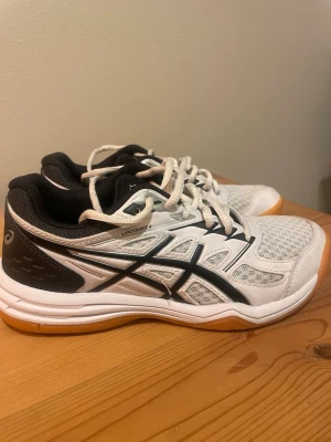 Vita Asics Upcourt 4 sneakers - Säljer ett par vita Asics Upcourt 4 sneakers med svarta detaljer och gummisula i orange. Skorna har meshpaneler för bra ventilation, klassisk snörning och låg profil. Perfekta för inomhussport eller träning. Snygg sportig look och bekväm passform.