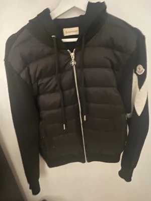 Svart Moncler pufferjacka med mönstrade ärmar - Säljer en svart pufferjacka från Moncler med huva och dragkedja. Jackan har stickade ärmar i svart och vitt med zigzag-mönster och Moncler-logga på ena ärmen. Kroppen är quiltad och vadderad, medan ärmarna är mjuka och stickade. Perfekt för dig som vill ha en unik och snygg jacka. Den är i storlek S men passar bäst för en som bär Xs. Tveka inte angående frågor bara att höra av sig!! Samt dustbag och tags följer med!