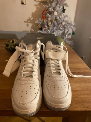 Nike Air Force 1 Hi Retro QS vita - Nike Air Force 1 Hi Retro QS i helvitt skinn med högt skaft, klassisk snörning och kardborreband runt ank soppen. Snygg retrostil med perforerad tå och robust sula. Perfekta för dig som gillar ikoniska sneakers med clean look.