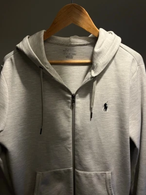Ralph lauren zip - Säljer en stilren ljusgrå hoodie från Ralph Lauren med dragkedja och huva. Tröjan har snörning i huvan och den klassiska svarta Polo-loggan broderad på bröstet. Perfekt för en avslappnad look. Jätte fint skick och inga slitningar förutom att de är lite nopprigt vid bröstet men syns knappt, skriv privat om du har några funderingar!