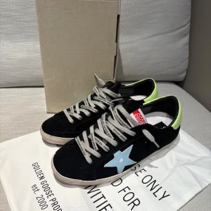 Golden Goose Superstar sneakers svart/blå - Golden Goose Superstar sneakers i svart mocka med ljusblå stjärna på sidan och limegröna detaljer vid hälen. Grå snören med logga och vit sula. Klassisk låg modell med rund tå och platt sula, perfekt för en cool och avslappnad stil.  Storlek 43. Vid fler funderingar och bilder.. HÖR av dig ;)