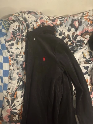 Svart skjorta med röd Polo Ralph Lauren märke - Säljer en svart  skjorta med röd ralph lauren märke från Polo Ralph Lauren, helt ny använd med klassisk krage och vita knappar. Skjortan har lång ärm och den ikoniska röda loggan broderad på bröstet. Perfekt för dig som gillar stilrena och griscg plagg.