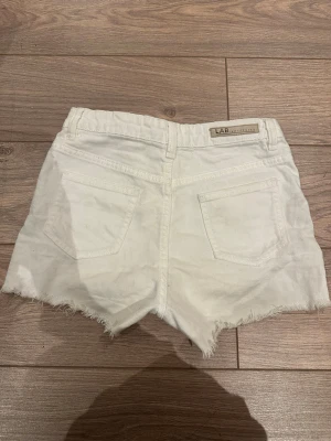Vita jeansshorts från LAB Industries - Snygga vita jeansshorts från LAB Industries med råa, fransiga kanter och klassisk femficksdesign. Shortsen har bälteshällor, dragkedja och knapp framtill. Perfekta för varma dagar och en chill look.