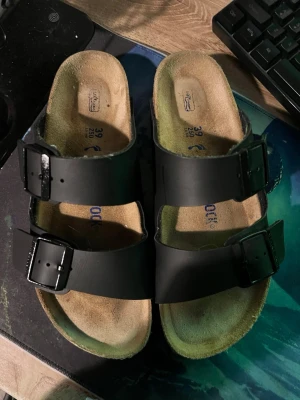 Svarta Birkenstock sandaler strl 39 - Säljer ett par klassiska Birkenstock sandaler i svart med två breda remmar och justerbara spännen. Ovandelen är i skinn och sulan är ergonomiskt utformad för extra komfort. Perfekta för sommaren och riktigt sköna att gå i.