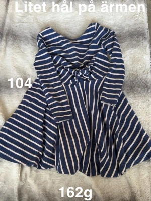 Blåvit randig klänning med knyt - Supersöt blåvit randig klänning med lång ärm och knyt i midjan. perfekt för dig som gillar en avslappnad men ändå stilren look. Passar till både chill och mer uppklädda tillfällen.🌸 Litet hål på ärmen. Storlek 104