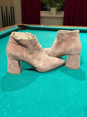 Beige mocka stövletter med klack - Säljer ett par snygga beige stövletter i mocka med spetsig tå och bred klack. Skorna har dragkedja på sidan för enkel på- och avtagning. Perfekta för dig som vill ha en stilren och trendig look till hösten eller vintern.