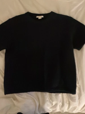 Svart kortärmad stickad topp - En enkel och stilren svart stickad topp med korta ärmar och rund hals. Toppen har en rak passform och är perfekt för dig som gillar minimalistisk stil. Materialet känns mjukt och lite tjockare, vilket gör den extra skön att bära.