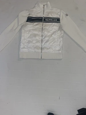 Vit Moncler cardigan jacka med dragkedja - Säljer en snygg vit cardigan jacka från Moncler med ribbstickade ärmar och nederkant. Framsidan har ett glansigt tyg med Moncler-logga och svart band över bröstet. Jackan har hög krage och hel dragkedja framtill. Perfekt för dig som gillar stilrena och exklusiva plagg.