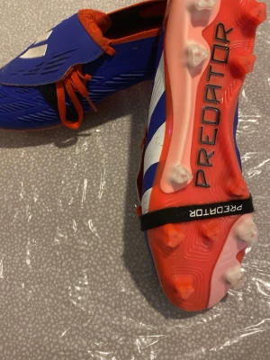 Adidas Predator blå fotbollsskor (FG) - Säljer ett par Adidas Predator fotbollsskor i blått med vita och orange detaljer. Skorna har snörning, täckt plös och dobbar för gräsplan. Tillverkade i syntetmaterial med texten 'HYBRIDTOUCH 2.0' på sidan och 'PREDATOR' under sulan.