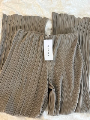 Beige plisserade byxor från Amisu - Säljer ett par beige plisserade byxor från Amisu med bred resår i midjan. Byxorna har ett loose fit och raka ben, tillverkade i ett mjukt syntetmaterial som ger en skön känsla. Perfekta för dig som gillar trendiga och bekväma plagg.