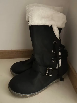 Svarta vinterstövlar med fuskpäls - Säljer ett par helt nya svarta stövlar med vit fuskpäls på insidan och upptill. Skorna har dekorativ snörning på sidan, spänne med metallbuckle och grov sula.