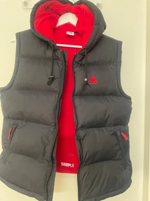 Svart dunväst M från Le Coq Sportif - Säljer en svart dunväst från Le Coq Sportif med röd broderad logga på bröstet. Västen har huva med dragsko, röda detaljer på dragkedjorna och en quiltad look. Perfekt för lager på lager under höst och vinter och tidig vår. 