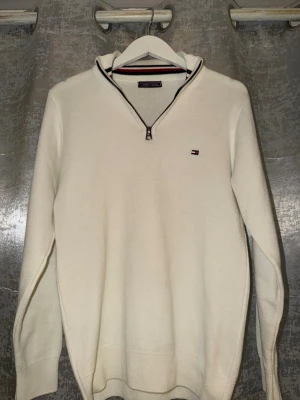  Tommy Hilfiger quarterback zip - Säljer en vit stickad tröja från Tommy Hilfiger med halv dragkedja och diskret logga på bröstet. Tröjan har lång ärm och ribbade muddar. Perfekt till lager på lager och enkel att matcha med jeans eller chinos.