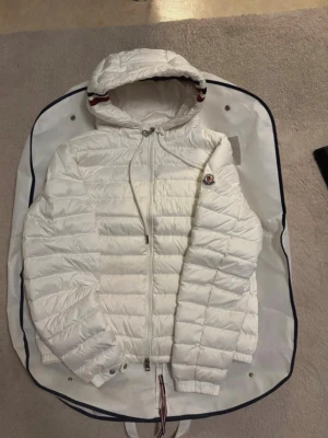 Vit Moncler pufferjacka med huva - Säljer en snygg vit pufferjacka från Moncler med huva och dragkedja framtill. Jackan har Moncler-logga på bröstet och coola rödblå detaljer på huvan. Perfekt för kalla dagar och riktigt stilren look.