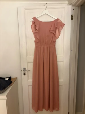 Lång rosa klänning från NLY Eve - Supersöt långklänning i ljusrosa från NLY Eve. Klänningen har volangdetaljer vid ärmarna, v-ringning i ryggen. Perfekt till bal eller liknande 