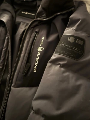 Svart pufferjacka från Sail Racing - Säljer en svart pufferjacka från Sail Racing med logga på bröstet och patch på ärmen. Jackan har huva, två bröstfickor med dragkedja och en clean, sportig look. Perfekt för kalla dagar och riktigt snygg till streetwear.