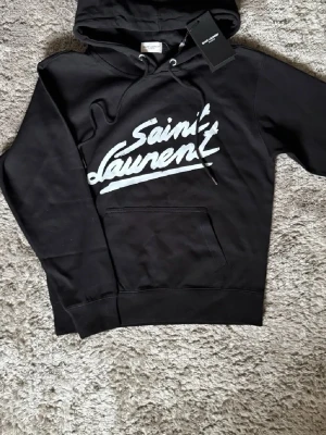 Saint Laurent  - Exklusiv hoodie från Saint Laurent i svart – en tidlös och lyxig piece med den ikoniska loggan framtill. Perfekt för en stilren och modern look med premiumkänsla. 🔺 Storlek S – slim passform ⚜️ Helt nytt skick med tagg 🔺 Tillverkad i mjuk bomull av hög kvalitet 📦 Fraktas inom 24 timmar & trygg affär