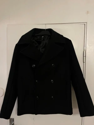 Svart skepparkavaj från H&M - Klassisk svart skepparkavaj från H&M med dubbelknäppning och bred krage. Jackan har raka ärmar och två sidofickor. Perfekt för dig som vill ha en stilren och tidlös look under höst och vinter.