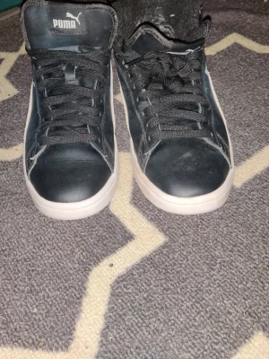 Svarta Puma sneakers med foder inuti  - Säljer ett par svarta Puma sneakers med vit sula och klassisk vit sidostrip. Skorna har snörning, SoftFoam+ innersula för extra komfort och en stilren design. Materialet är en mix av syntet och mocka, perfekt för dig som gillar sportig streetstyle.