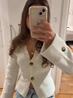 Vit kofta med knappar - Säljer en stilren vit kofta med stora dekorativa knappar framtill. Koftan har v-ringning, lång ärm och en figurnära passform. Perfekt att matcha med jeans för en snygg look. Använd 1 gång.