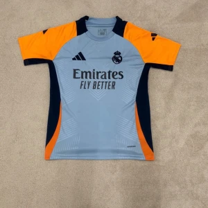 Real Madrid Adidas träningströja - Snygg Real Madrid träningströja från Adidas i ljusblått med marinblå och orange detaljer. Tröjan har korta ärmar, klubbmärke och Adidas-logga på bröstet samt 'Emirates Fly Better' tryckt framtill. Perfekt för fotbollsträning eller att bära till vardags.