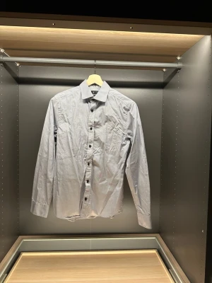 Randig skjorta från Riley Slim Fit - Snygg randig skjorta från Riley i slim fit-modell. Skjortan är vit med tunna mörkblå ränder, har långa ärmar och klassisk krage. Tillverkad i bomull och stängs med mörka knappar framtill. Perfekt för dig som gillar stilrena detaljer.