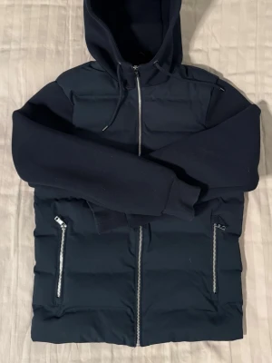 Next hybrid jacka - Snygg mörkblå pufferjacka med huva och dragkedja framtill. Jackan har quiltad front, släta ärmar och två fickor med dragkedja. Perfekt för kyliga dagar. Såldes ut snabbt. 