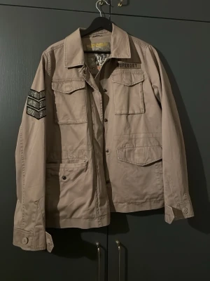 Beige overshirt från Superdry med paljetter - Säljer en grön/beige jacka från Superdry med stora fickor fram och detaljer på ärmen och på ryggen. Jackan har klassisk krage, knappar framtill samt dragkedja och en relaxed passform. Använd 1-2 gånger