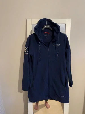 Race Marine blå hoodie, storlek S - Mörkblå långärmad hoodie med dragkedja från Race Marine. Jackan har dragsko i huvan och två fickor med dragkedja fram. En siffra 83 är tryckt på vänster ärm.