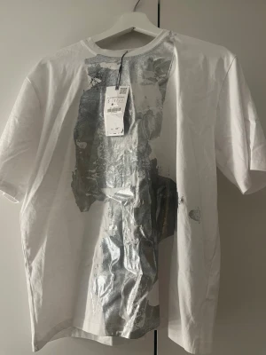 Vit t-shirt med silvertryck från Zara - Säljer en vit t-shirt från Zara i storlek S med ett stort abstrakt silverfärgat tryck framtill. T-shirten har klassisk passform, rund hals och korta ärmar. Materialet är mjuk bomull som känns skönt mot huden. Perfekt för dig som gillar statement-plagg.