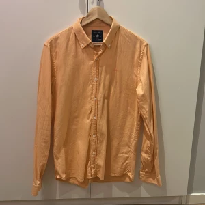 Aprikosfärgad skjorta från Robbie Moor. - Persikofärgad skjorta från Robbie Moor i slim fit-modell. Klassisk button-down krage, vita knappar framtill och lång ärm. Skjortan är tillverkad i bomull och har en diskret broderad logga på bröstet. Perfekt för dig som gillar stilrena plagg med snygg passform. Skicka vid minsta lilla intresse🤩