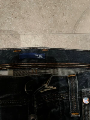 G-Star RAW 3301 mörkblå jeans - Säljer ett par mörkblå G-Star RAW 3301 jeans med snygga slitningar och kontrastsömmar. Klassisk femficksmodell med tryckt logga på myntfickan och raka ben. Jeansen har en cool tvättad look och är tillverkade i robust denim.