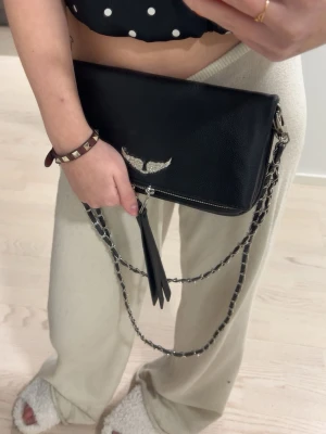 Svart Zadig & Voltaire axelväska i skinn - Säljer en svart axelväska från Zadig & Voltaire i skinn med silverfärgad kedja och snyggt emblem med strass framtill. Väskan har två dekorativa tofsar och dragkedja upptill. Perfekt storlek för det viktigaste och ger en cool vibe till outfiten.