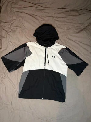 Under Armour jacka - Snygg vindjacka från Under Armour i svart, grått och vitt. Jackan har huva med snörning, dragkedja framtill och färgblockad design. Loggan sitter på bröstet och materialet är lätt och syntetiskt, perfekt för aktiva dagar. Skriv om ni har frågor!