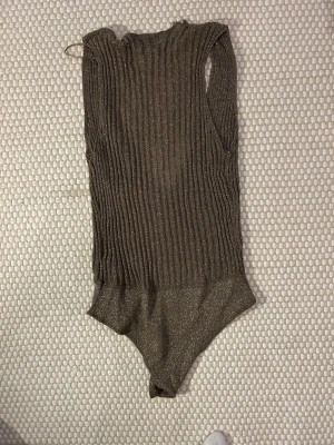 Glittrig body från Zara - Säljer en glittrig guldig body  från Zara med omlott och djup v-ringning. Västen är ribbad och har en croppad passform som sitter snyggt till jeans eller kjol. Materialet är mjukt och stretchigt med metallictrådar för extra shine. Se hål, varav priset! Kan enkel sys igen.