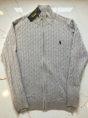 Ljusblå kabelstickad zipper kofta från Polo Ralph Lauren - Snygg ljusblå kabelstickad kofta från Polo Ralph Lauren med dragkedja framtill och hög krage. Klassisk logga broderad på bröstet och ribbade muddar vid ärmslut och nederkant. Perfekt för lager på lager och stilren look.