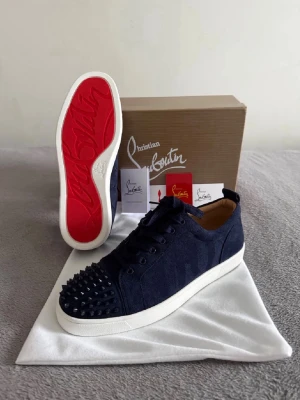 Mörkblå sneakers från Christian Louboutin - Säljer ett par mörkblå sneakers från Christian Louboutin med klassisk röd sula och svarta nitar på tån. Skorna har snörning, vit platt sula och är tillverkade i mocka för en lyxig känsla. Perfekta för dig som vill sticka ut med exklusiva detaljer.