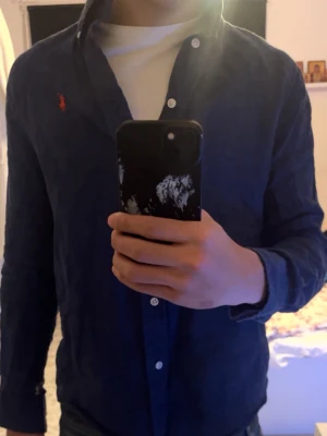 Ralph Lauren skjorta - Klassisk mörkblå linne skjorta från Ralph Lauren i storlek XS. Skjortan har långa ärmar, knappar framtill och den ikoniska röda broderade loggan på bröstet. Tillverkad i mjuk bomull för en skön känsla och snygg passform.