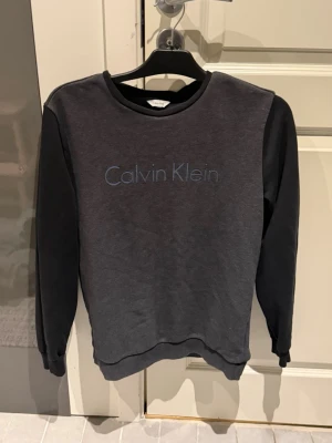 Mörkgrå sweatshirt från Calvin Klein - Säljer en stilren mörkgrå sweatshirt från Calvin Klein med svart logga framtill. Tröjan har svarta ärmar, rund halsringning och är tillverkad i mjuk bomullsblandning. Perfekt för dig som gillar enkel och clean stil.