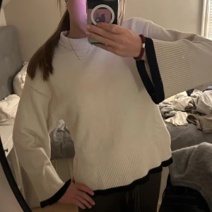 Vit stickad tröja  - Vit stickad tröja från Gina Tricot med svarta detaljer längs ärmslut och nederkant. Tröjan har breda, långa ärmar och ribbade muddar samt en högre ribbad halsringning. Perfekt för dig som gillar en clean och stilren look.